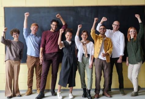 A group of eight adults cheerfully standing with raised fists in front of a blackboard indoors.