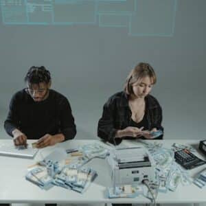 Two people counting money at a desk with computers and cash equipment.