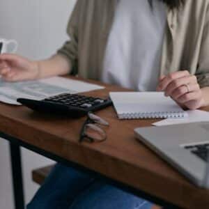 A woman manages finances at home, using a laptop and calculator on a wooden desk.