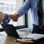 Crop unrecognizable coworkers in formal wear standing at table with laptop and documents while greeting each other before meeting