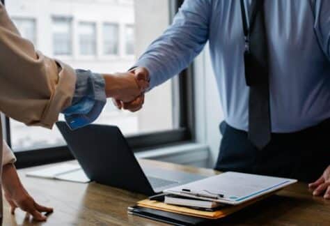 Crop unrecognizable coworkers in formal wear standing at table with laptop and documents while greeting each other before meeting