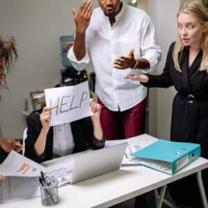 A group of employees in heated discussion, one holding a 'HELP' sign, indicates workplace stress.