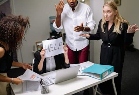 A group of employees in heated discussion, one holding a 'HELP' sign, indicates workplace stress.