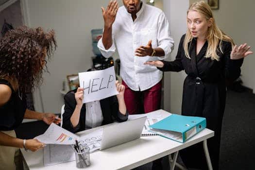 A group of employees in heated discussion, one holding a 'HELP' sign, indicates workplace stress.