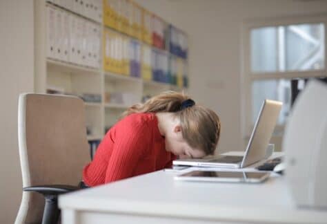 A tired woman in a red sweater leans her head on a desk with a laptop, symbolizing workplace fatigue.