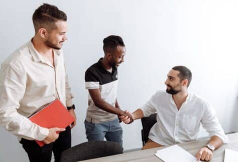 Three men in an office setting, shaking hands, representing teamwork and diversity.